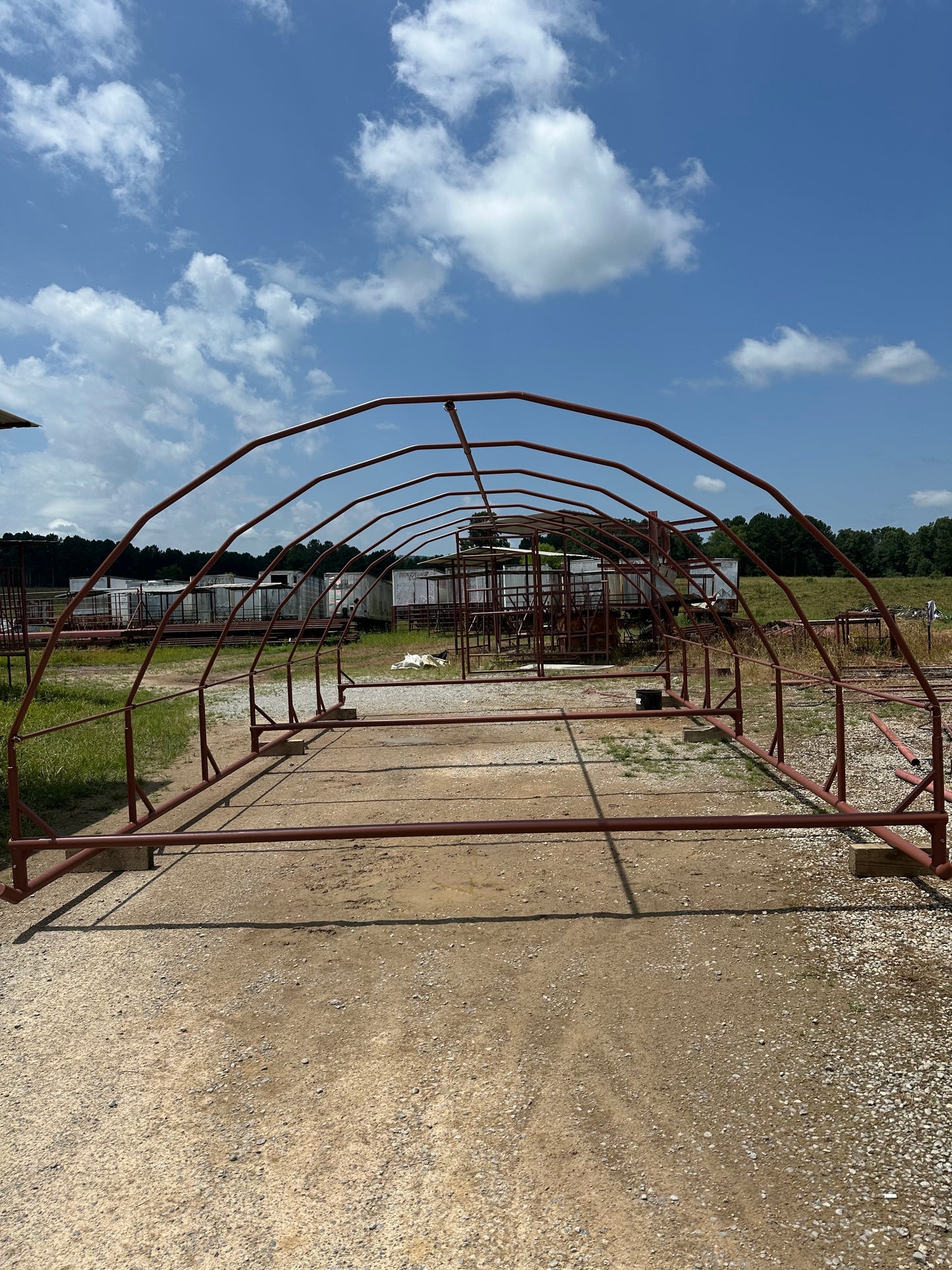 Red-Oxide-colored arched steel structure on a gravel surface with a blue sky and clouds in the background